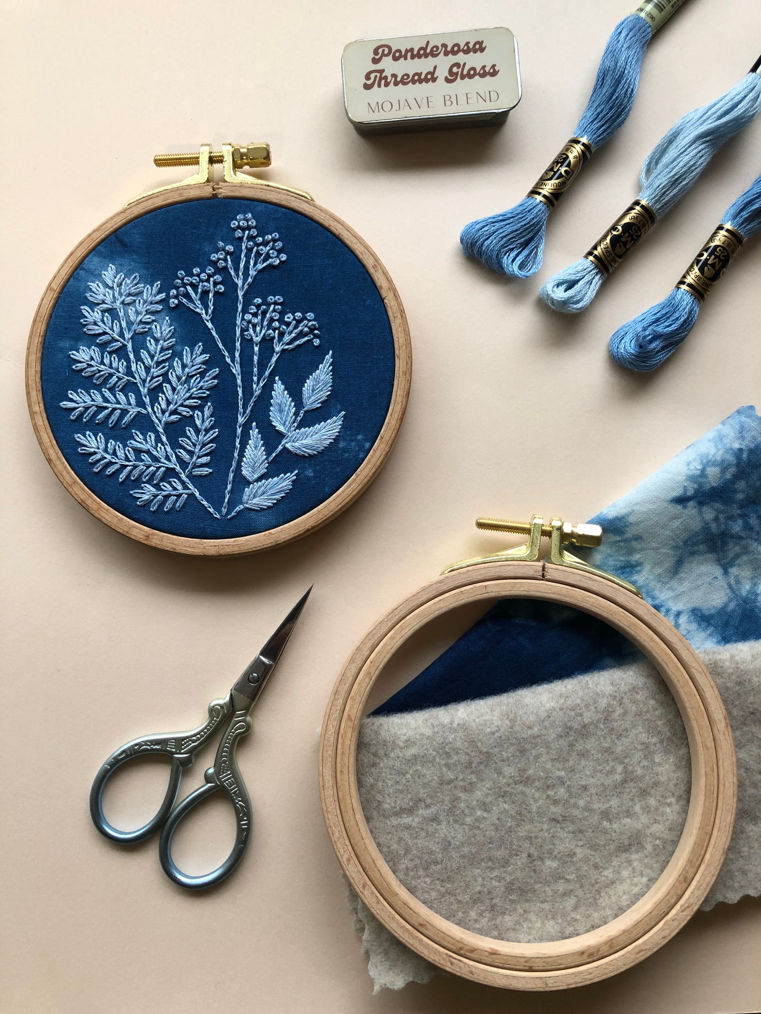 Some contents of the Cyanotype botanicals beginner friendly DIY embroidery kit are displayed: the finished design in a hoop,  indigo cotton fabric, a 4-inch beechwood embroidery hoop, 3 skeins of embroidery thread, felt, thread gloss, and embroidery scissors (not included in kit)