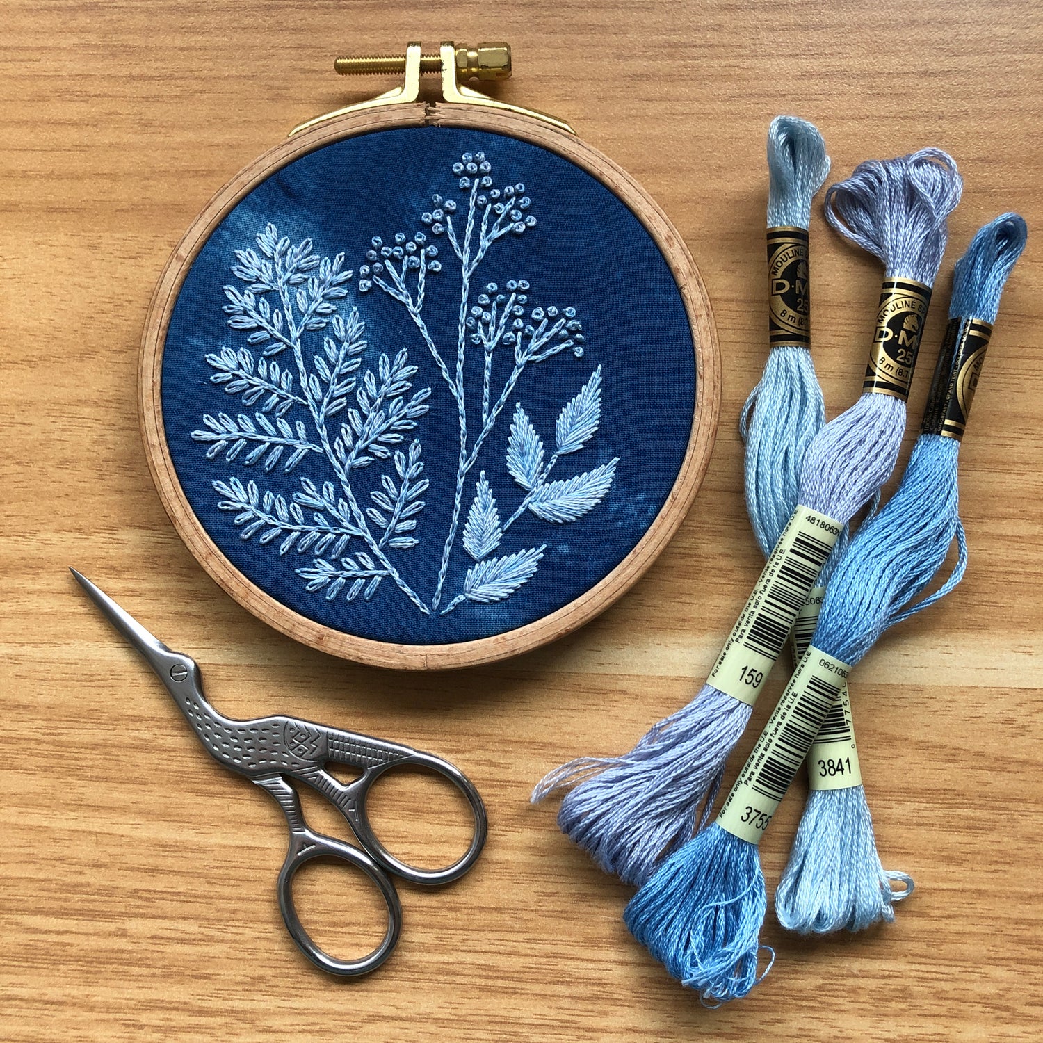 Completed Cyanotype botanicals design still in the embroidery hoop from the DIY embroidery kit. To the right are 2 skeins of thread, and to the left are embroidery scissors (not included in kit) 