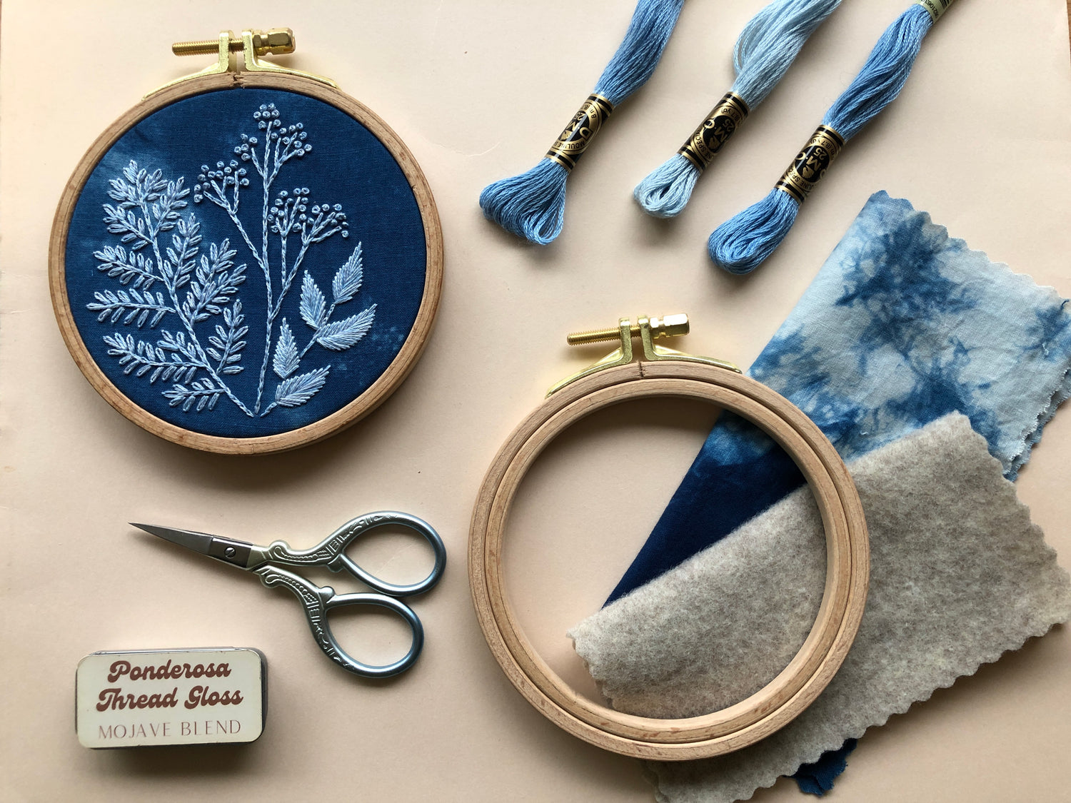 Some of the contents of the Cyanotype botanicals beginner friendly DIY embroidery kit laid out. A hoop with the finished design, embroidery scissors, 3 colors of thread, felt and fabric, a hoop, and thread gloss.