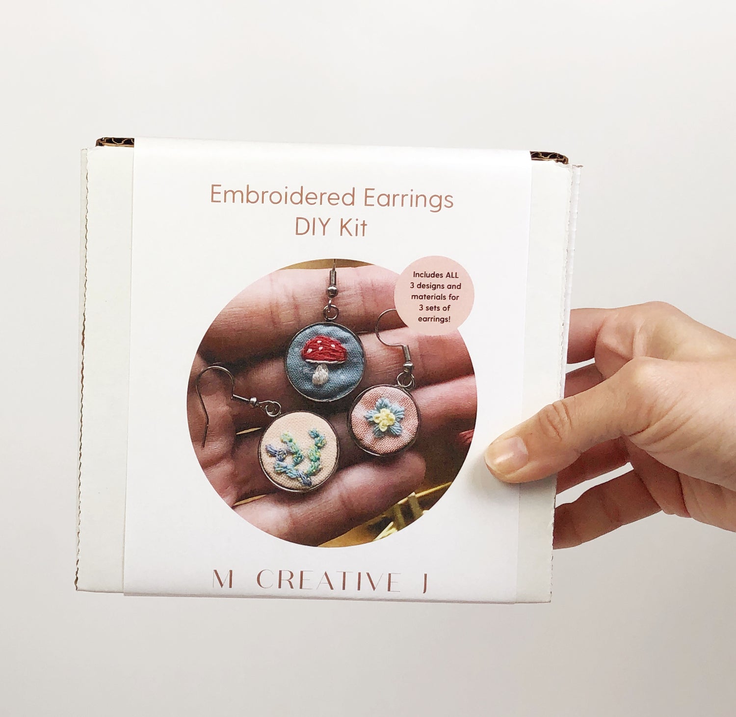 boxed DIY kit labeled 'Embroidered Earrings DIY Kit.' The kit features three designs of embroidered earrings, including floral and mushroom patterns, and includes materials for making three pairs of earrings