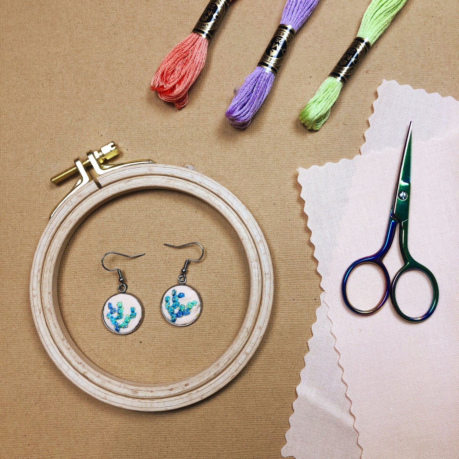 Two completed floral earrings from the beginner DIY embroidery earrings kit sit inside an embroidery hoop. 3 colors of thread are above and a pair of scissors to the right.