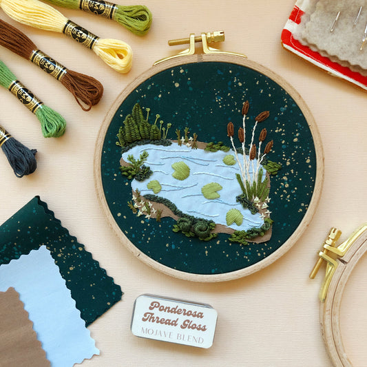 Contents of the 3D Lily Pad Pond advanced embroidery kit displayed: 5 skeins of thread, completed lily pad design still in the hoop, felt, thread gloss, sewing pins.