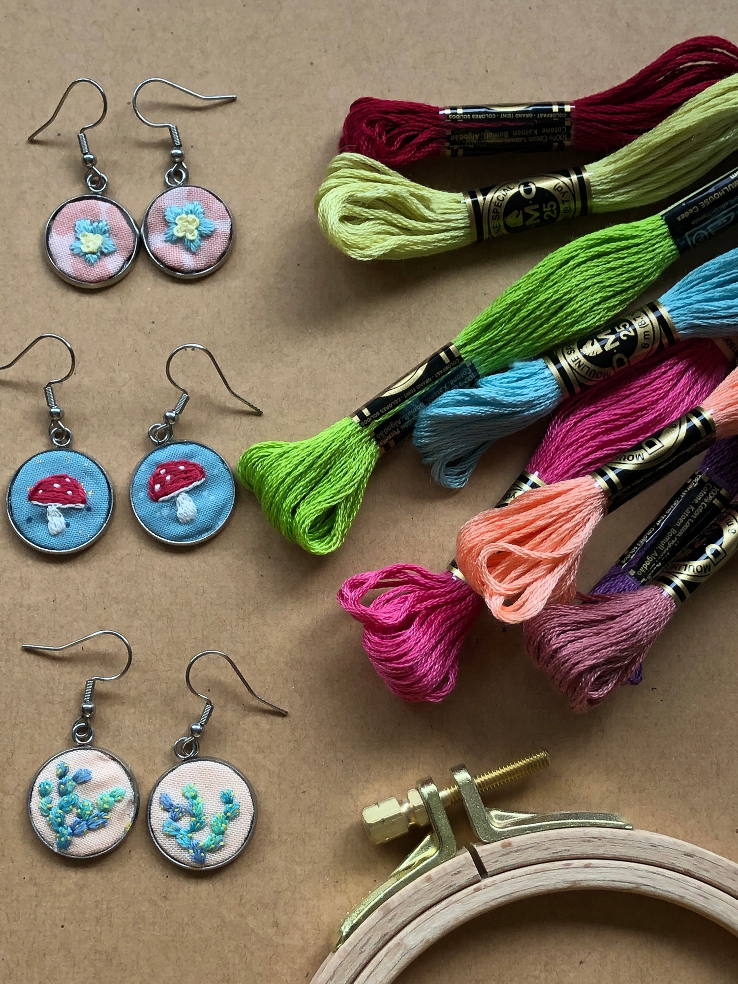 3 pairs of embroidered earrings with floral and mushroom designs from the earrings kit displayed next to the hoop and thread.