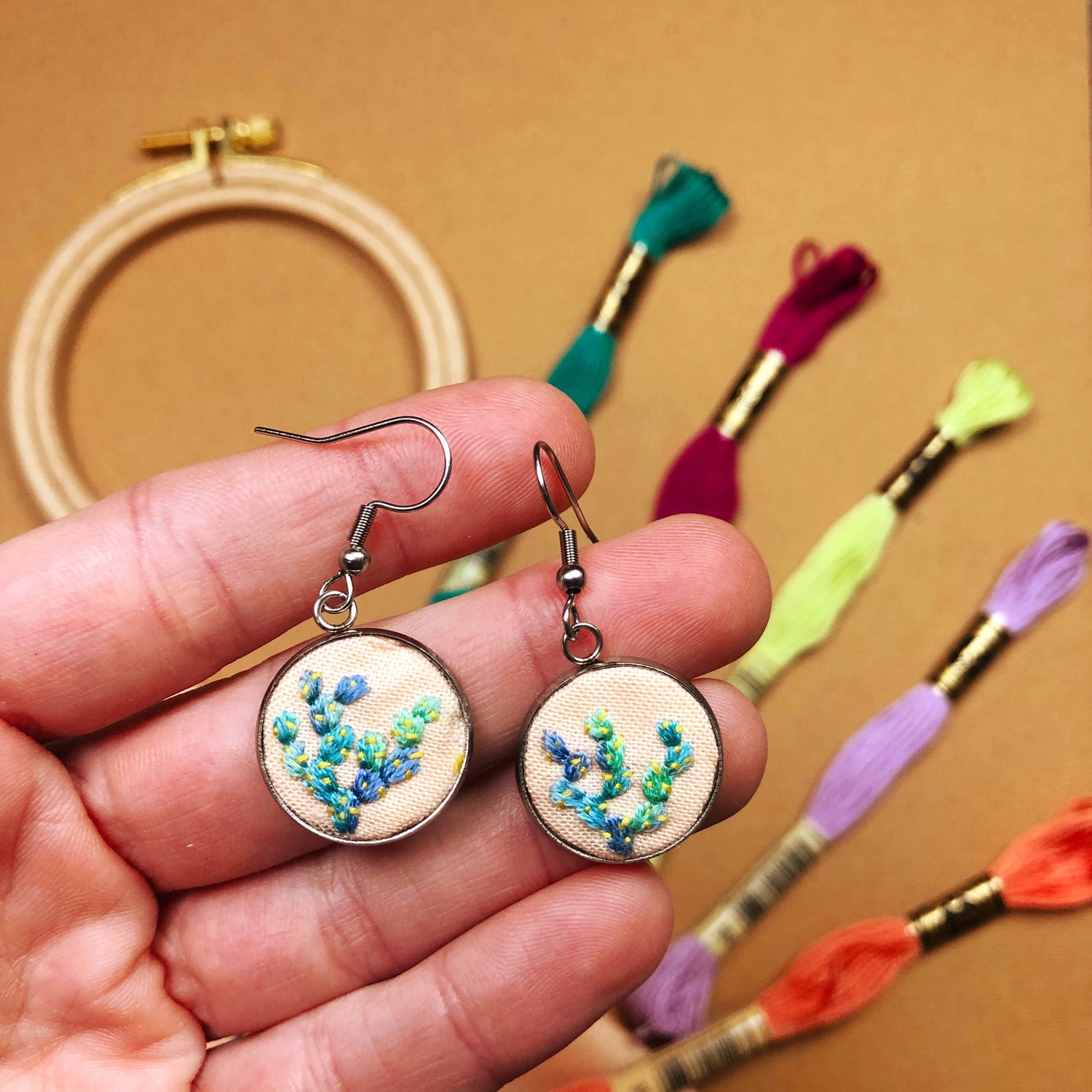 A hand holding completed embroidered earrings from the DIY beginner embroidery earrings kit. The hoop and thread is in the background.
