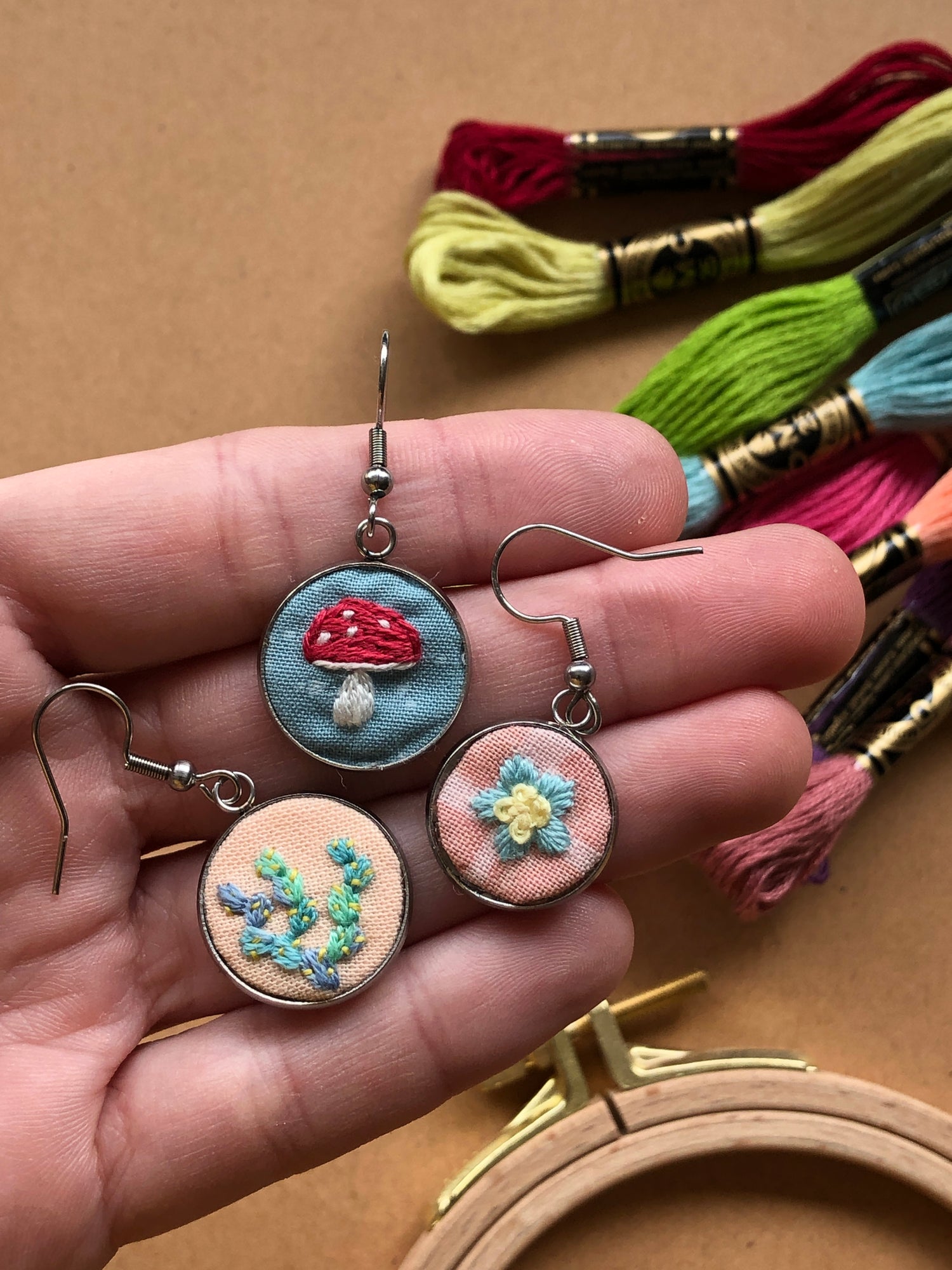 3 embroidered earrings with floral and mushroom designs sitting in the palm of a hand